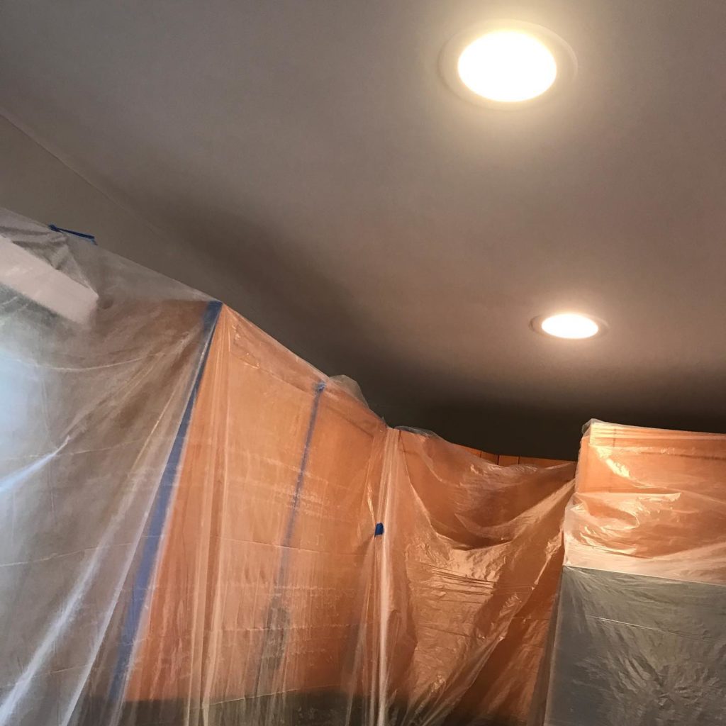 Room prepared for renovation or painting, with plastic sheets covering walls and furniture under recessed ceiling lights.
